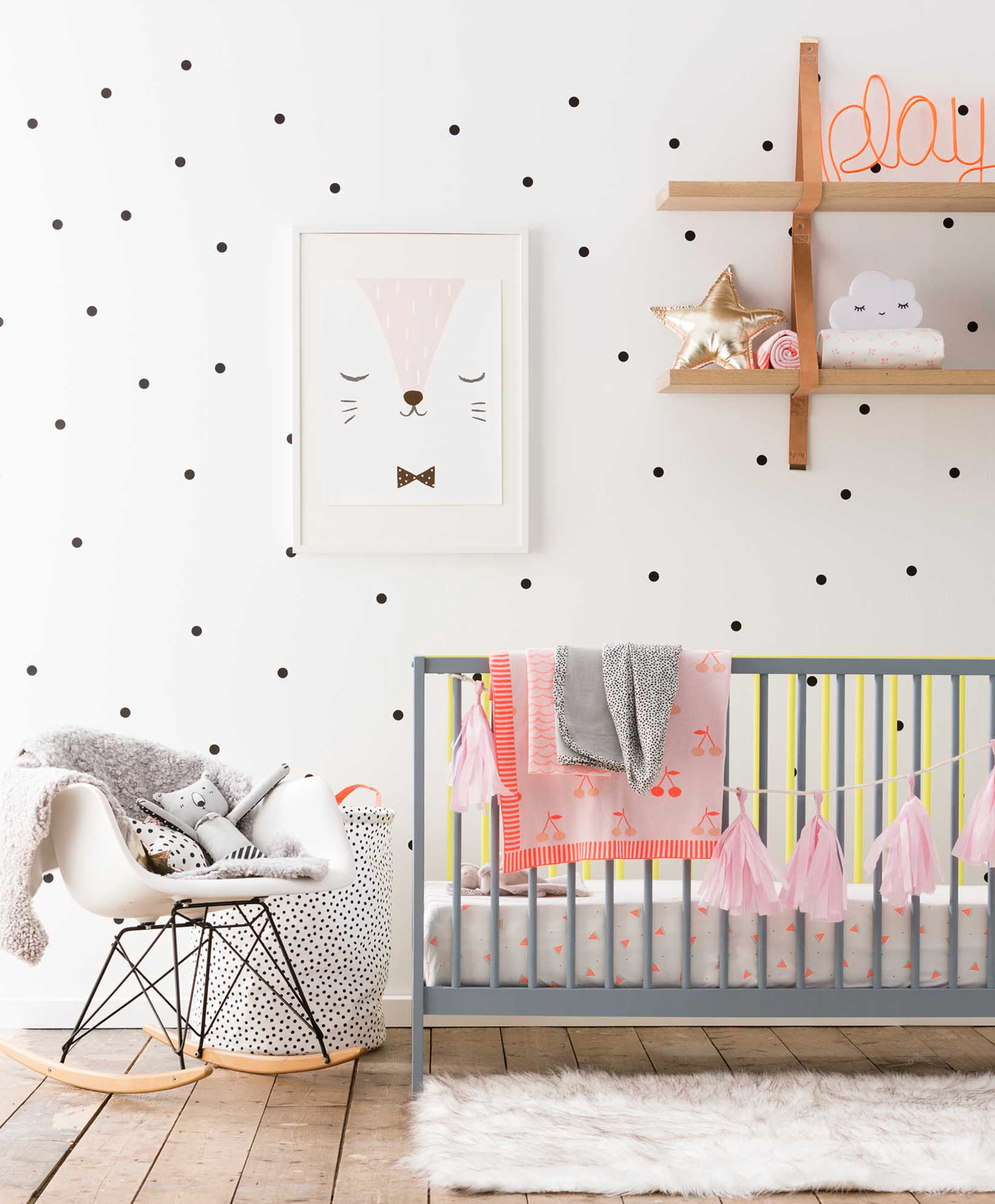 Small black polka dots on the nursery wall, grey crib, white Eames rocking chair and light brown wooden shelves